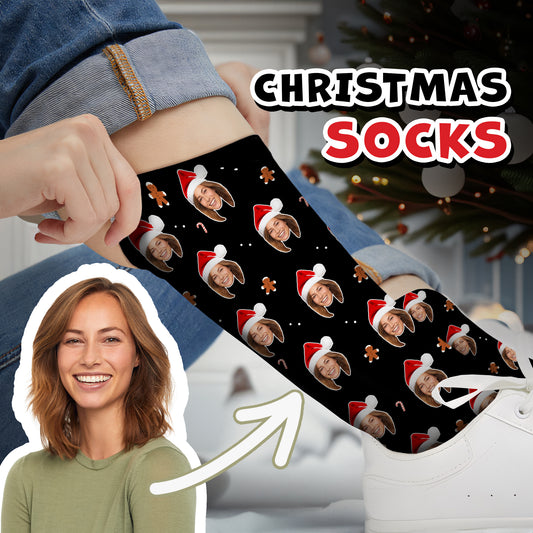 Christmas-themed personalized socks featuring a custom face in a Santa hat, surrounded by festive gingerbread men and candy cane patterns. The person is adjusting the black socks while sitting near a Christmas tree.